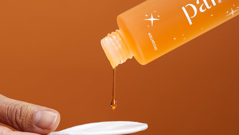 Close up image of a bottle of orange skincare liquid, Pai Bright Now Exfoliating Toner, being poured onto a reusable cotton pad